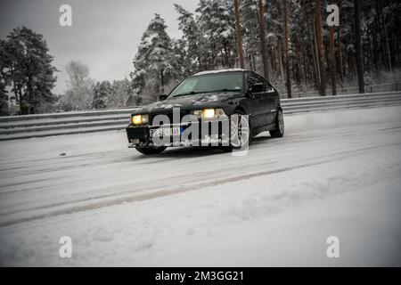 12-12-2022 Riga, Latvia  a black car driving down a snowy road next to a forest of trees and a fence with snow on it. . Stock Photo
