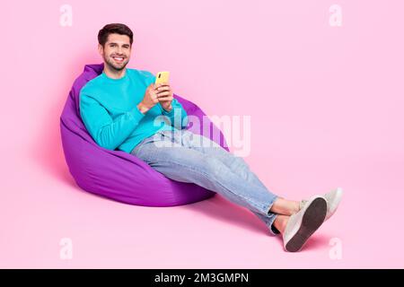Full body photo of nice young guy sitting hold device relax chatting read news wear trendy blue clothes isolated on pink color background Stock Photo