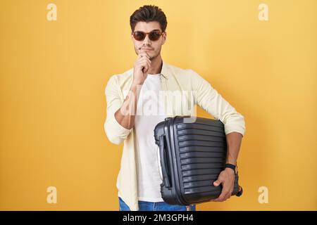 Young hispanic man holding suitcase going on summer vacation thinking worried about a question, concerned and nervous with hand on chin Stock Photo