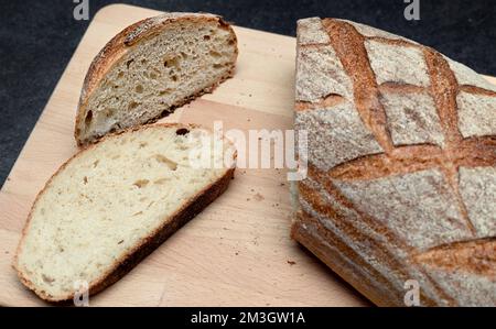 Oval loaf of freshly baked sourdough slided bread knife on cutting wooden board, angle view. Rustic sourdough bread on dark gray table Stock Photo