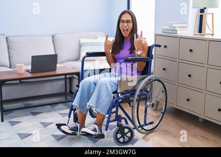 Young hispanic woman sitting on wheelchair at home shouting with crazy expression doing rock symbol with hands up. music star. heavy concept. Stock Photo