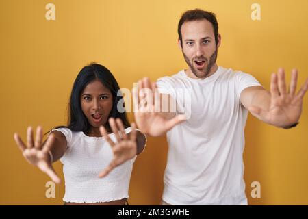 Interracial couple standing over yellow background doing stop gesture with hands palms, angry and frustration expression Stock Photo