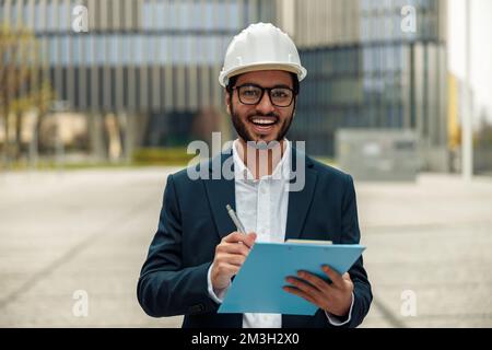 Indian construction architect in suit and hardhat with paper folder in hands against modern building Stock Photo
