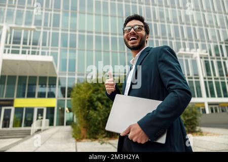 Smiling indian businessman in suit and glasses with laptop near office building Stock Photo
