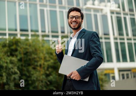 Smiling indian businessman in suit and glasses with laptop near office building showing thumb up Stock Photo