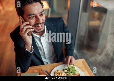 Happy indian businessman is talking phone with client during lunch time in cozy cafe Stock Photo