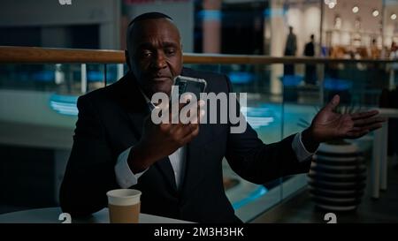 Ethnic African American man employer entrepreneur businessman in cafe with coffee tea talking business video call with mobile phone talk conference Stock Photo