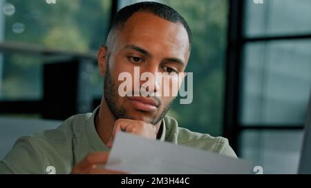 Calm African Latin guy man student entrepreneur open envelope notification from bank test result invitation reading news in paper admission letter at Stock Photo
