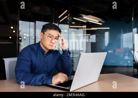 Serious thinking asian businessman working inside modern office, mature man in shirt and glasses using laptop at work, investor pondering complex decision. Stock Photo