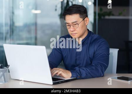 Serious thinking asian businessman working inside modern office, mature man in shirt and glasses using laptop at work, investor pondering complex decision. Stock Photo