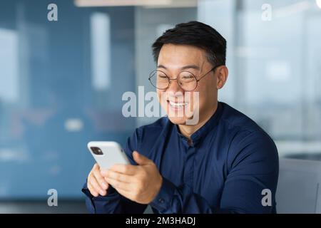 Cheerful and smiling asian businessman inside office using phone, man typing message and browsing online pages and watching video, boss in glasses and shirt at work. Stock Photo
