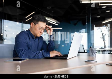 Asian boss working inside office, man overtired with headache, businessman in shirt and glasses using laptop at work, sitting at desk at workplace. Stock Photo