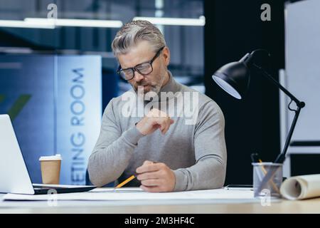 Concentrated senior male architect working in the evening by a lamp in the office. Sitting at the table, drawing a plan with a pencil, working with papers. Stock Photo
