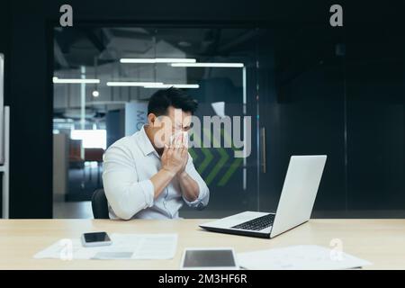 Illness at work. A young Asian man sits in the office at the table, wipes his nose with a napkin, has a runny nose, flu, feels bad, is exhausted. Stock Photo