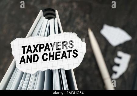 On the table is a notebook on which lies a piece of torn paper with the inscription - Taxpayers and cheats. The pen lies outside the sharpness zone. Stock Photo