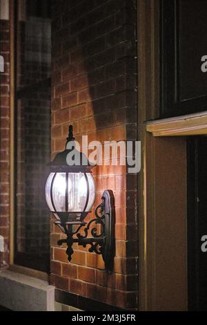 An old decorative black metal lamp on a brick walled building illuminates the dark nighttime shadows. Stock Photo