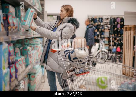 The concept of shopping and parenting. A young mother chooses baby food with her baby sitting in a grocery cart. Close up. The concept of family shopp Stock Photo