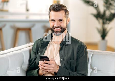 Happy joyful handsome caucasian bearded man, sit at home on a sofa in living room, uses his smart phone, relaxing on couch, messaging online in social media, browsing internet, looks at camera, smiles Stock Photo