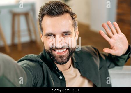 Cheerful attractive positive caucasian bearded man, in a casual shirt, takes a selfie on his smartphone, waves hand, looks into the front camera of the mobile phone, smiles friendly Stock Photo