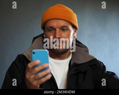 Man at home sitting on sofa, using smartphone at cold with winter coat and yellow hat. Stock Photo