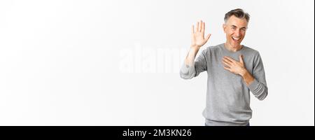 Portrait of handsome middle-aged man making promise, raising one hand and swearing to tell truth, smiling at camera, standing over white background Stock Photo