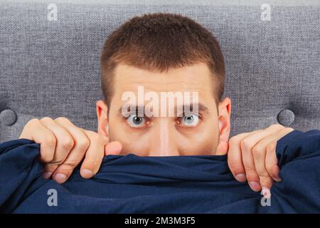 Looking straight into the frame. A handsome young guy hiding behind a blanket looks to the side. Direction of gaze in different directions. Stock Photo