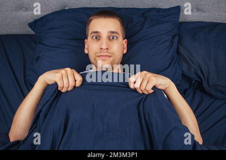 Looking straight into the frame. A handsome young guy hiding behind a blanket looks to the side. Direction of gaze in different directions. Stock Photo