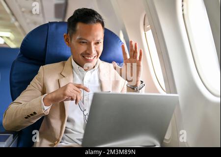 Professional and successful Asian businessman or male CEO waving his hand while having an online meeting or conference with a client during the flight Stock Photo