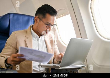 Stressed and busy Asian businessman managing his business tasks, reviewing financial report and using laptop during the flight. Stock Photo