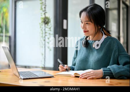 Smart and pretty young Asian female college student concentrating on her homework, using laptop computer and doing homework on school book. Stock Photo