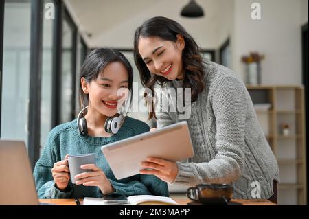 Attractive and cheerful Asian female office worker showing something on tablet screen to her colleague, enjoy chatting on the coffee break in the offi Stock Photo