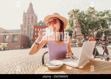 A girl drinks coffee in an old European city of Cologne and works or studies on a laptop. Remote employment and student life Stock Photo