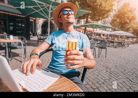 Happy man relaxes and drinks a glass of beer in a pub in the fresh air and works or communicates via laptop Stock Photo