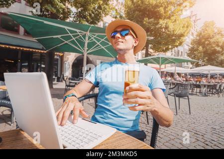 Happy man relaxes and drinks a glass of beer in a pub in the fresh air and works or communicates via laptop Stock Photo