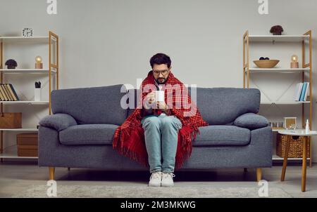 Sick man in a blanket sitting on the couch at home, freezing and drinking hot tea Stock Photo