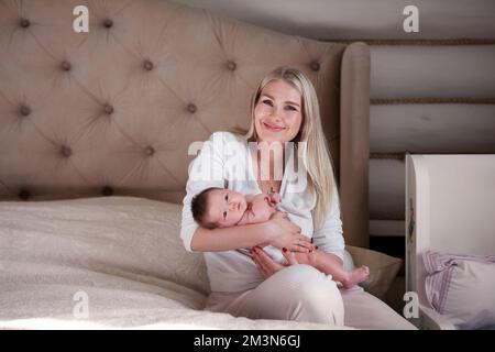 Happy mother holds newborn baby in her arms, smiles sitting on bed and looks at camera. Beautiful blonde mom with long hair and her child at home Stock Photo