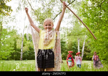 Portrait of happy girl with hands raised holding stick standing in grass by friends at park Stock Photo