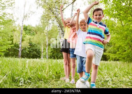 Portrait of smiling children with hands raised on grass in park during summer holiday Stock Photo