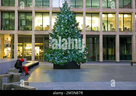 Person wearing red sitting on bench by Christmas tree office building in winter New Street Square London EC4 UK England Great Britain  KATHY DEWITT Stock Photo