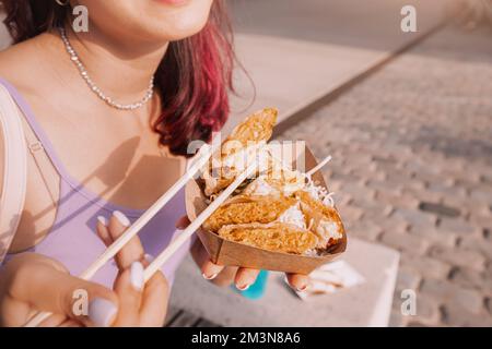 Happy girl eating spring rolls from takeaway paper box using chopsticks at city street. Fast food eatery and asian cuisine Stock Photo