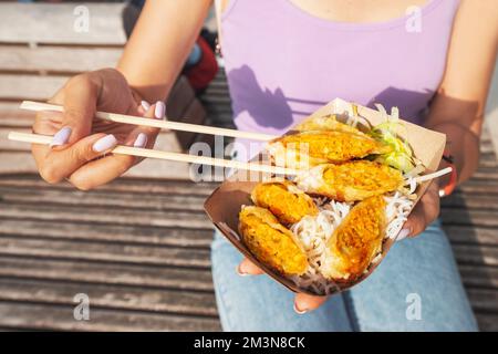 Happy girl eating spring rolls from takeaway paper box using chopsticks at city street. Fast food eatery and asian cuisine Stock Photo