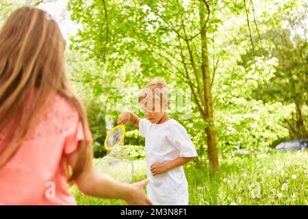 Boy playing with soap bubbles by female friend in green garden during summer vacation Stock Photo
