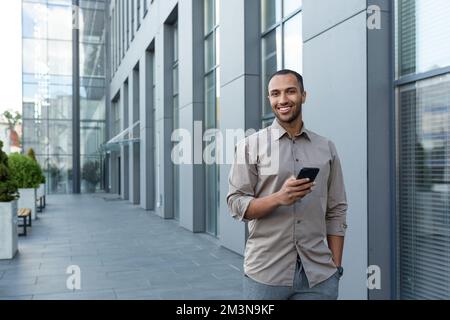Hispanic businessman walking outside office building, man holding smartphone, smiling and looking at camera. Stock Photo