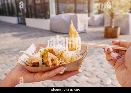 Happy girl eating spring rolls from takeaway paper box using chopsticks at city street. Fast food eatery and asian cuisine Stock Photo