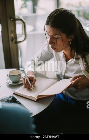 Beautiful young college student writing something in her notebook at the cafe bar with friends Stock Photo