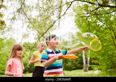 Boy enjoying while making soap bubbles by female friends in park during summer vacation Stock Photo