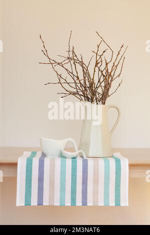 Stylish minimalistic still life, dishes on striped tablecloth or napkins with jug with branches on wooden table against beige wall background Stock Photo