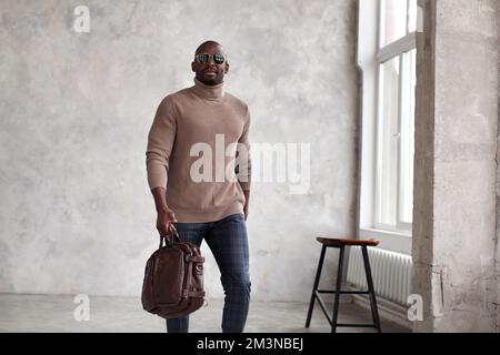 Handsome african american man walking indoors wearing fashionable clothes beige sweater, trousers and leather bag, sunglasses Stock Photo