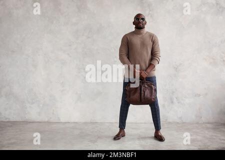 Fashion man stands against gray background, wearing stylish clothes, a turtleneck sweater, plaid trousers, brown boots loafers, bag and glasses Stock Photo