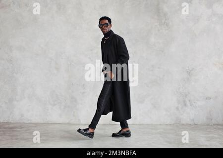 Fashionable man wearing black coat, turtleneck, pants, loafers shoes, glasses in studio. Fashion african american male model in stylish clothes, total Stock Photo